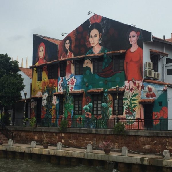 A building painted with street art of 4 different ethnically diverse women on the edge of the Malacca River