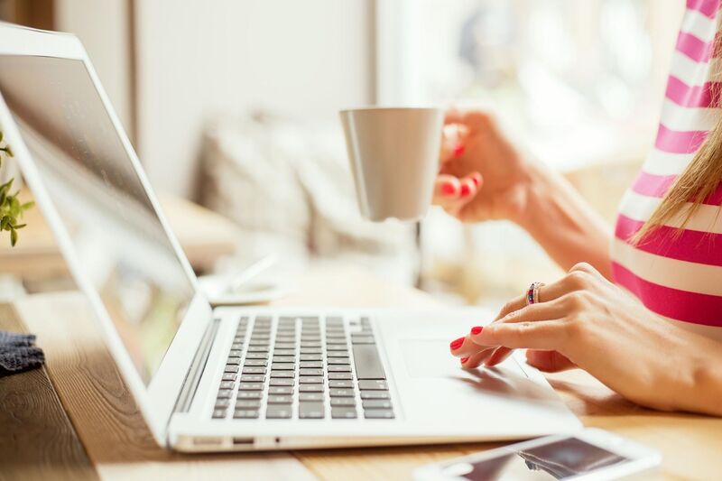 Woman typing on her laptop with her left hand with a cup of coffee in her right hand. She is wearing a red and white striped tshirt