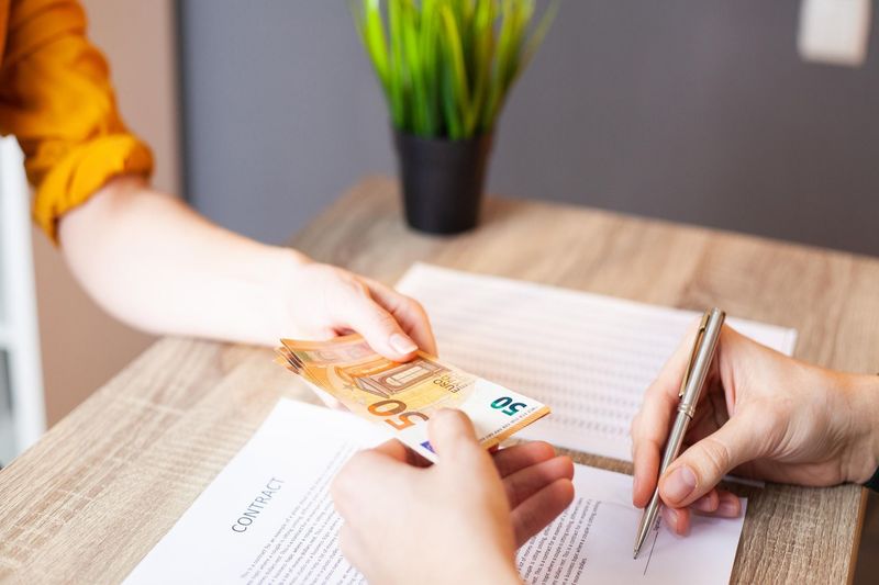 two people handing Euro currency to each other ovr a desk while one person signs a contract.