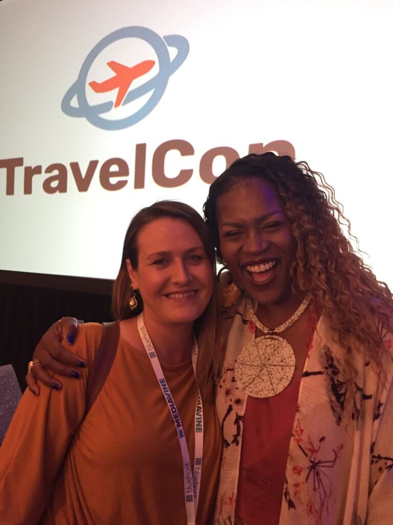 a personal photo of myself wearing a yellow sweater and my event lanyard, being hugged by Gloria Atanmo. she is wearing a red top, white jacket and a white beaded necklace. they are standing in front of a sign that says travelcon. the name of the travel conference