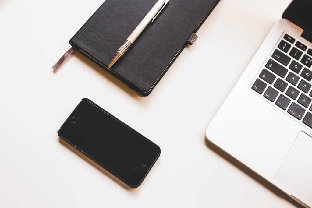 a black iPhone next to a notebook with a pen on top and an open laptop in the frame to the left sitting on a table at a travel conference