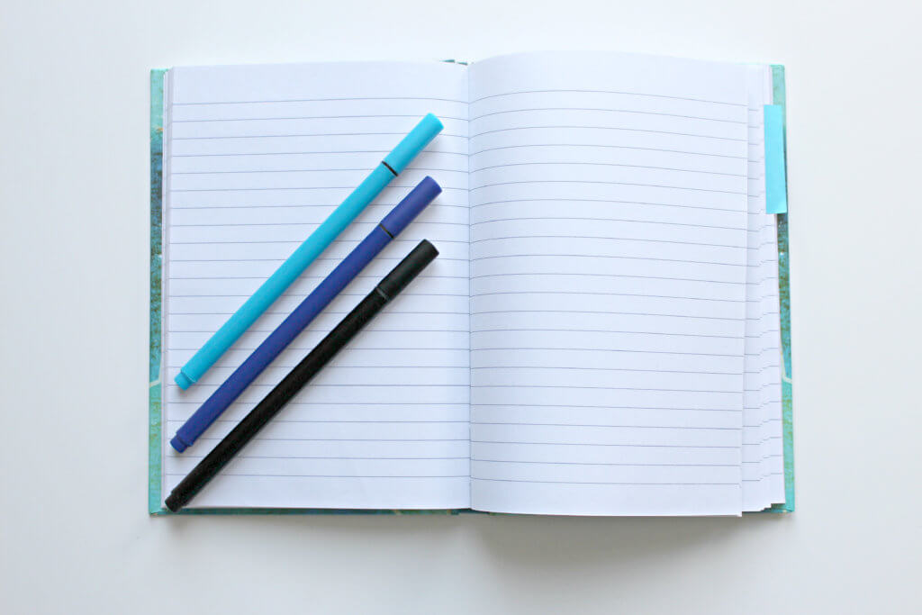 a light blue, dark blue and black pen laying a top an open notebook