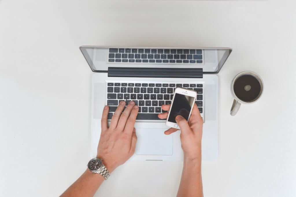 a man has an open laptop, he is typing with his left hand and holding his iPhone with his right. there is a black coffee sitting to the right of the computer. he wears a silver watch on his left wrist 