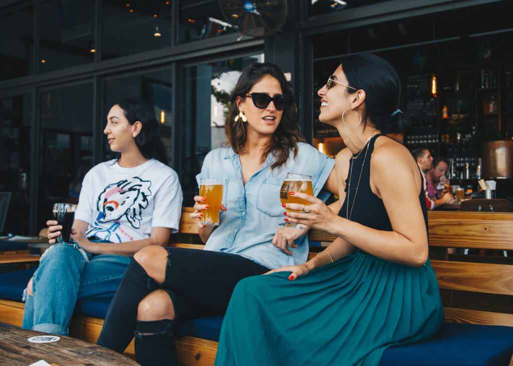 three women holding beer glasses are networking and talking at a bar. they are siting on a long bench