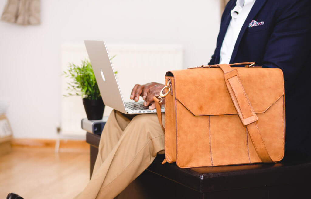 a man sitting down with a laptop on his lap at a travel conference. The laptop is open and he is typing. he is wearing a blue blazer, white shirt and tan pants. his legs are crossed at the ankle and he has a tan satchel sitting next to him