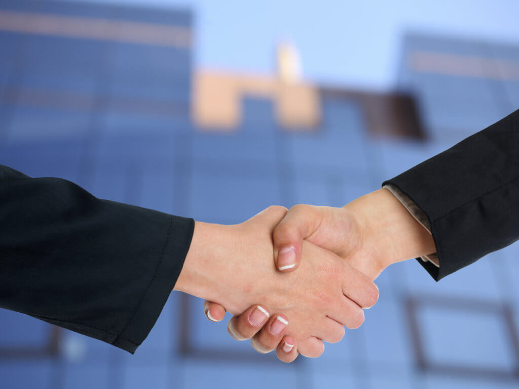 two females, shaking hands at a travel conference. both wearing black blazers visible from the forearm down to the manicured nails