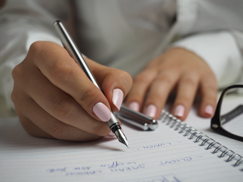 a woman is writing notes in an open notebook in blue pen. she is wearing a white shirt and has manicure pink nails. her pen lid is sitting on the book and her glasses are slightly noticeable to the left of the image