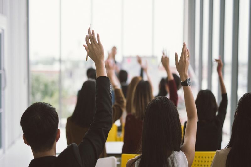 a grup of people in a room facing forward with their hands raised as if wanting to ask a question