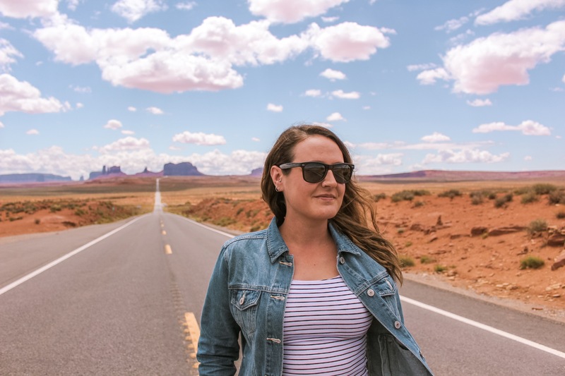 Luisa standing in the middle of the highway at Monument Valley. She is wearing a while and black stiped top, denim jacket and black RayBan sunglasses. she is facing the camera but looking to the right of the photo. she is a solo traveller