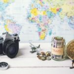 passport, notebook, camera, model air plane, money jar labelled travel with some USD inside and coins sitting adjacent to the jar. A straw hat and sunglasses all sitting a top a table and all in front of a world map