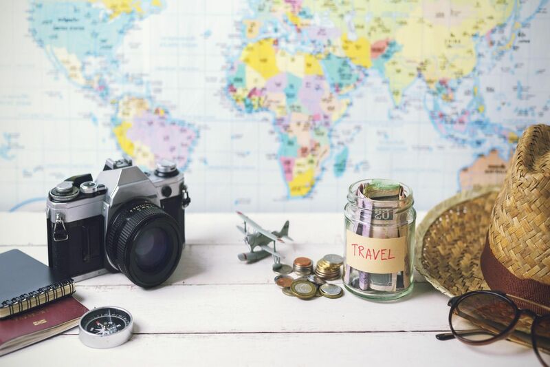 passport, notebook, camera, model air plane, money jar labelled travel with some USD inside and coins sitting adjacent to the jar. A straw hat and sunglasses all sitting a top a table and all in front of a world map