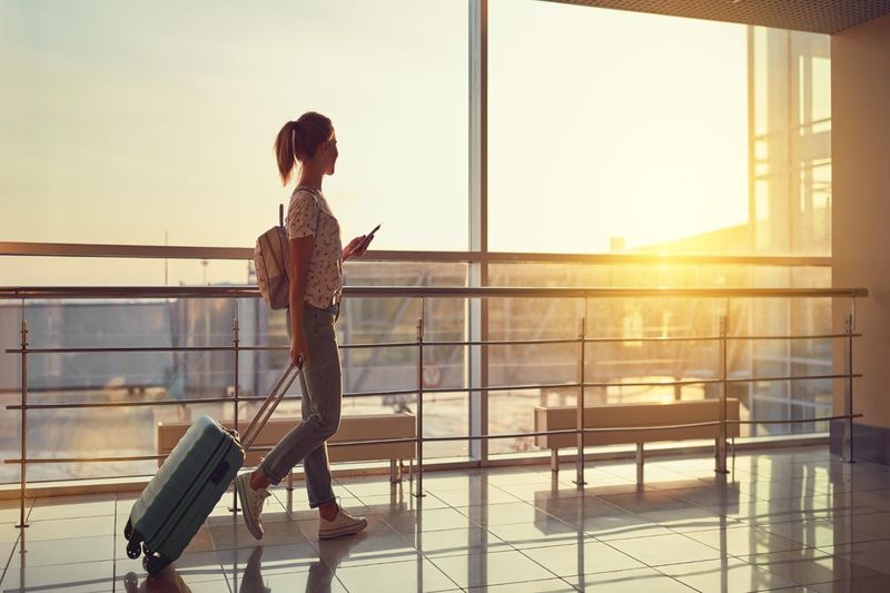 woman walking through the airport. She is dragging a big piece of luggage and has a small backpack on her back. She is holding her phone up and slightly in front of her. The sun is golden and setting through the window