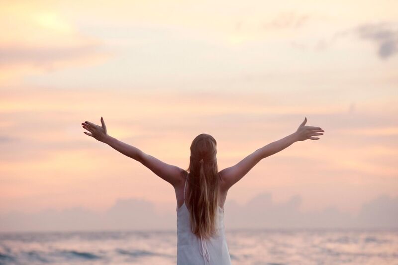 woman facing away from the camera toward the beach at sunset with arms outstretched