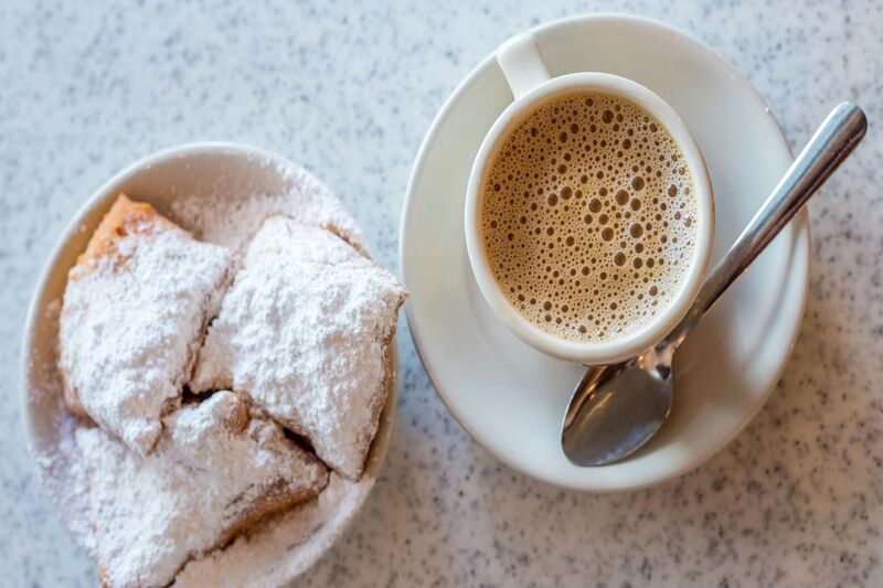 a plate of 3 Beignets on the left with a cup of chicory coffee on the right, on a saucer with a teaspoon