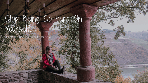 a woman sits on a low wall between two pillars. she is looking off into the distance down to a river. she is wearing a red jacket, black jeans and black shows. er blonde hair is down around her shoulders