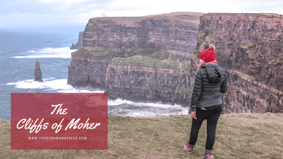 a woman stand on  huge cliff, looking over the ocean at another cliff. she is wearing black jeans and black jackets and a red fluffy beanie with a brown bauble on top