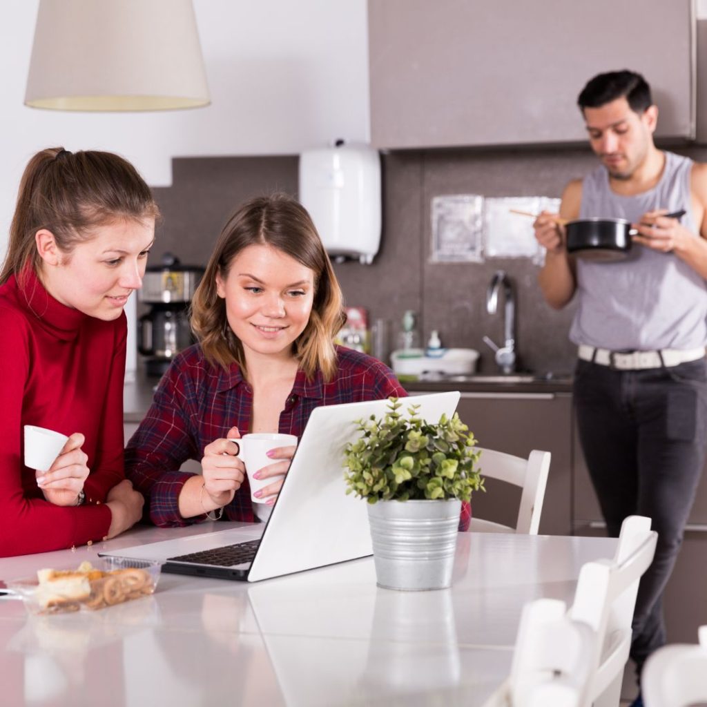 group of travellers in hostel common room. Two friends on a laptop at the table and a make guest in the background eating food straight from the saucepan