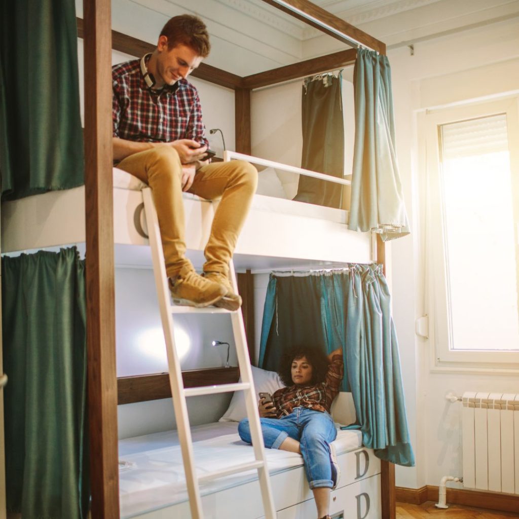 two friends enjoying hostel life on their bunk beds. a lady lounging on the bottom bunk reading and the a man hanging over the top bunk resting his legs on the ladder watching his phone