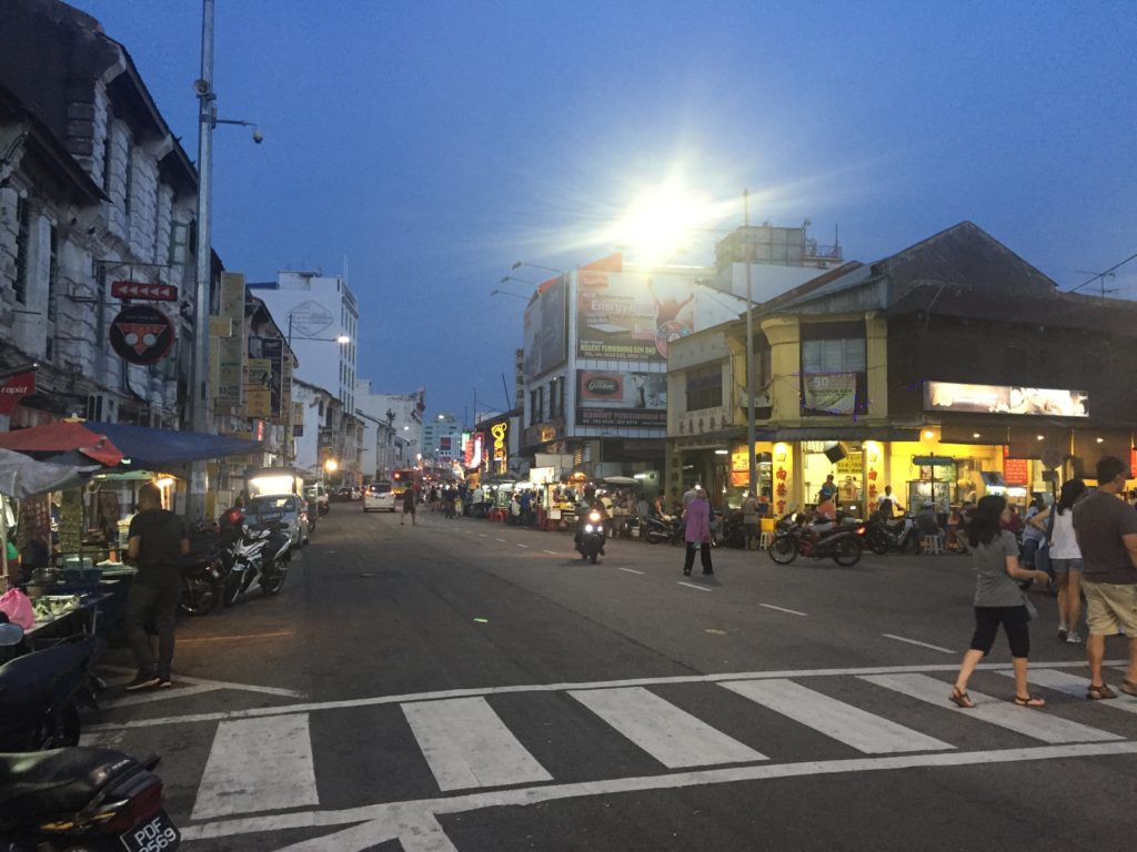 A night time scene of Love Lane in Penang, Malaysia. People are crossing the street to the restaurant. other people are riding their motorbikes down the street