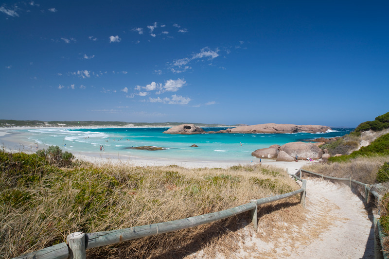 a walkway leads down to the protected bay of Twilight Bay. on either side of the path is sand dunes and vegetation. the beach sweeps around to the right. there are granite boulders out in the water