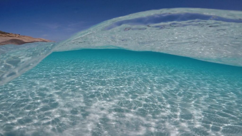 the photographer has taken this photo whilst swimming in the blue water. she has captured a wave curling over the lens. you can see under and over the wave with a small portion of teh shorelin above the curling wave to the left of the image