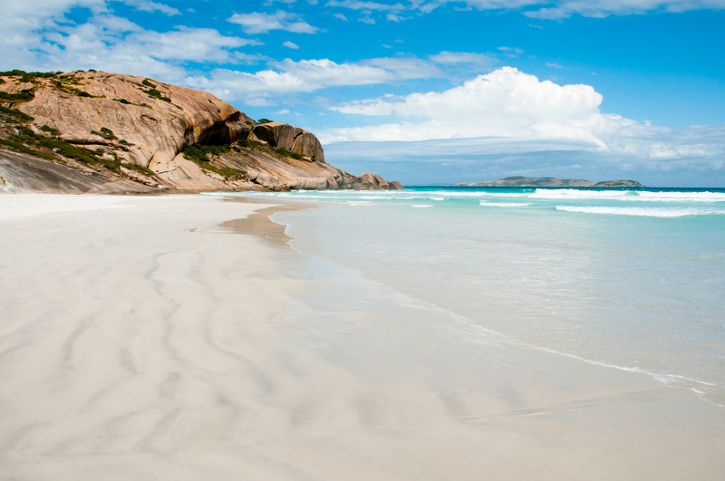 a long stretch of white sandy beach, with the blue water of the ocean to the right of the image. A rocky shoreline can be seen in the distance protruding into the water and cutting off the beach