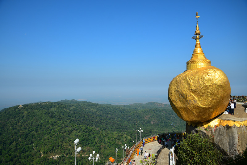 the golden boulder with a small golden pagoda teeters on the edge of a cliff to the right of the image. below the boulder to the left is a walk way with people gazing at the site. off into the distance is dense rainforest