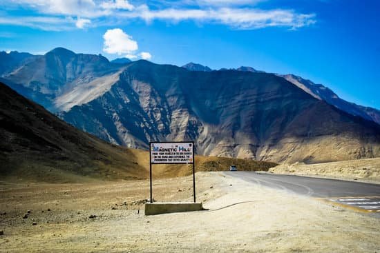 a view of magnetic hill, with the marked spot on the road to the left of the image and signage referring to magnetic hill in the centre. huge towering hills are in the background set against a blue sky with a streak of clouds from left to right in the sky