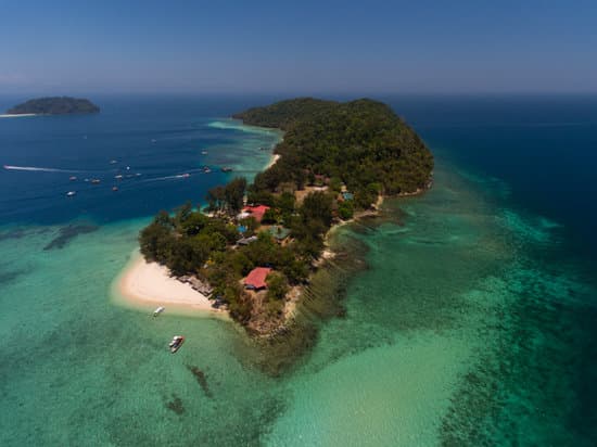 Aerial view of an island in Tunku Abdul Rahman marine Park. the island is surrounded by beautiful clear water. on the island are a few buildings with red roofs. the majority of the island is dense rainforest. there are a few boats anchored around the island. there is another smaller island in the distance to the left of the large island. A truly tropical Asia Landmark