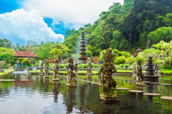 Tirta Gangga Water park. statues and round stepping stones are set in the tranquil water. the sky is blue yet some fluffy white clouds linger. to the left is a large rainforest covered mountain overlooking this beautiful Asia Landmark