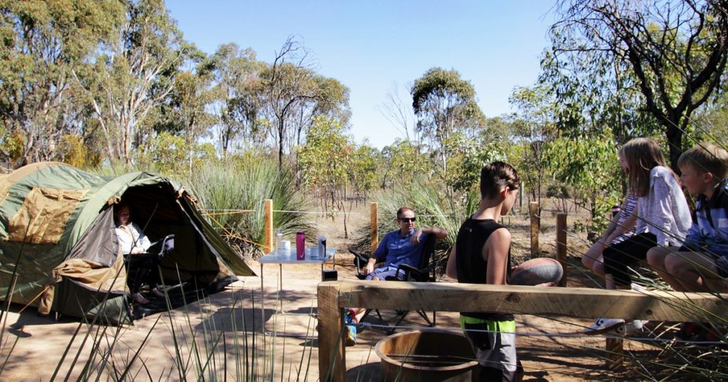 a group of campers are sitting in chairs around their tent at Wirra Campsite in northern Adelaide. the campsite is in low scrubby bushland native to Australia