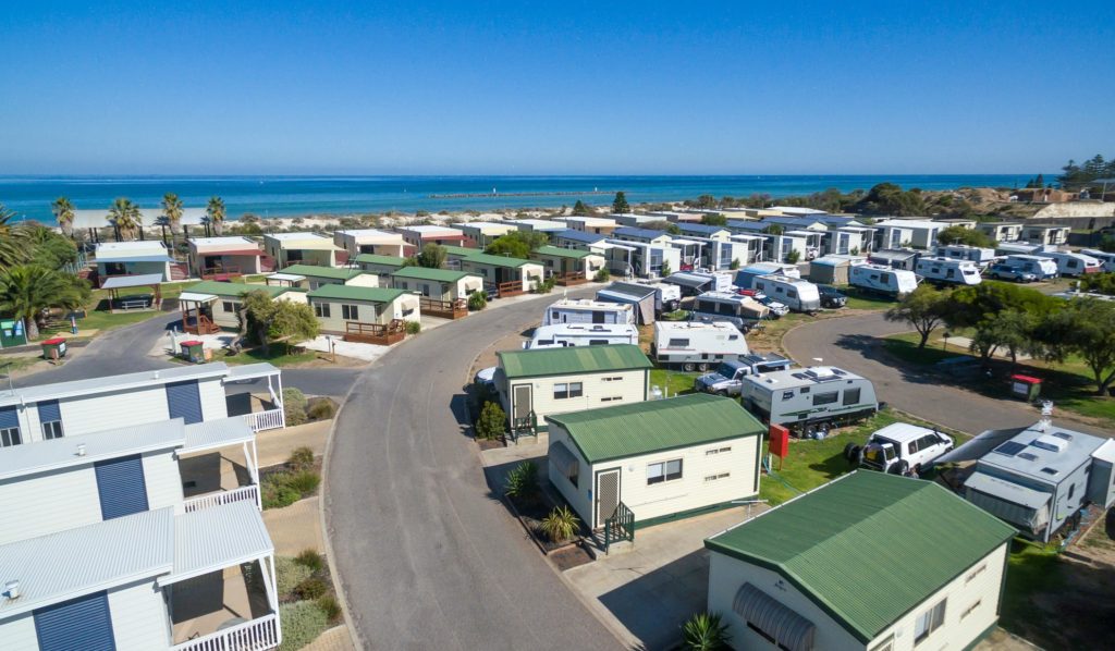 an aerial view of Adelaide Beachfront Semaphore caravan park. there is a road that sweeps to the right and it is surrounded by cabins and caravans. the ocean can be seen in the background