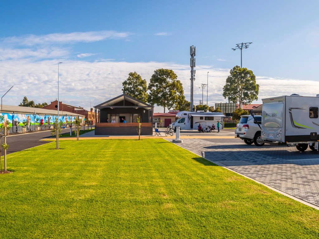 two caravans and four wheel drive car are parked on the pavement with a beautiful green patch of grass leaing from teh foreground of the image to the background. in the background is the camp kitchen for the caravan aprk