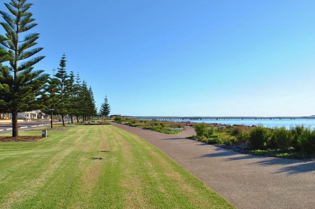 the blue ocean is on the right of the image with a footpath running along its edge. the grassy green surface of the campground is on the left of the image next to the footpath. there is a very long jetty off in the distance