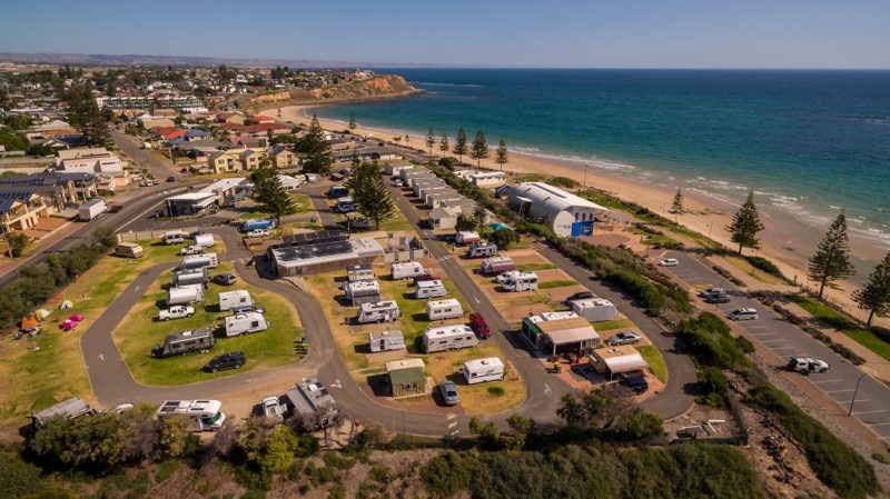 an aerial view of Christies Beach Holiday park. there are numerous cars abd caravans parked at the sites within the grounds. the beach is to the left and the distinct orange cliffs of the Fleurieu peninsula can be seen in the distance