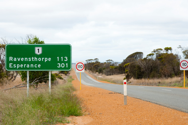 highway to Esperance surrounded by red dirt on both sides and with a green road sign saying the kilometres left to travel to Ravensthorpe and Esperance. there is also a speed sign indicating the maximum speed on the highway is one hundred and ten kilometres per hour