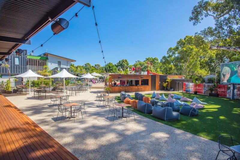 the image shows the outdoor cinema area of the marion holiday Park. there are beanbags spread out on the grass, an outdoor bar, umbrellas and tables and chairs on the pavement. the apartment accommodation can be seen in the distant