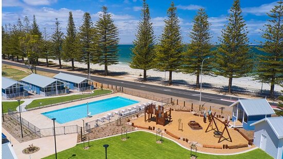 an aerial view of RAC Esperance holiday and caravan park. In the image is a grassy area in the foreground, a blue swimming pool, a playground. there is a road in the distance lined by tall pine trees and the sand and beach beyond the pines