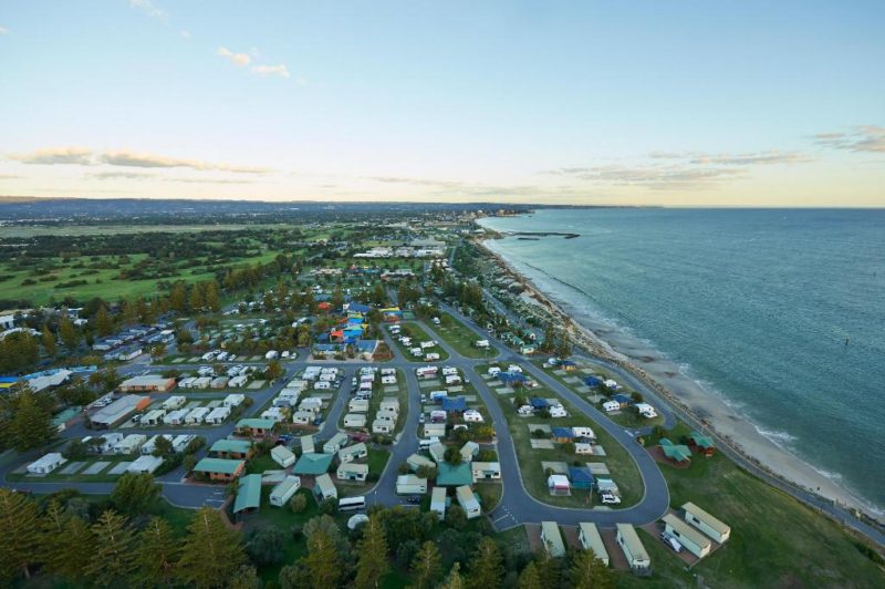 an aerial view of West Beach caravan park. the caravan park is right on the shores of the beach to the right and surrounded by green grass, shay trees and green fields to the left