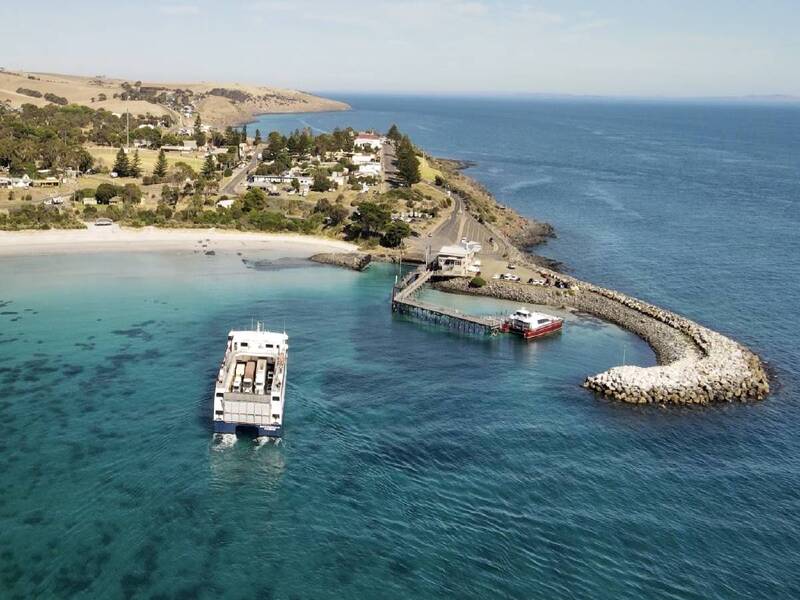 The Sealink ferry is pulling into the Penneshaw ferry terminal on Kangaroo Island. The water is clear, yet blue. There is a breakwater to the right of the image and a road leading from the terminal past houses in Penneshaw in