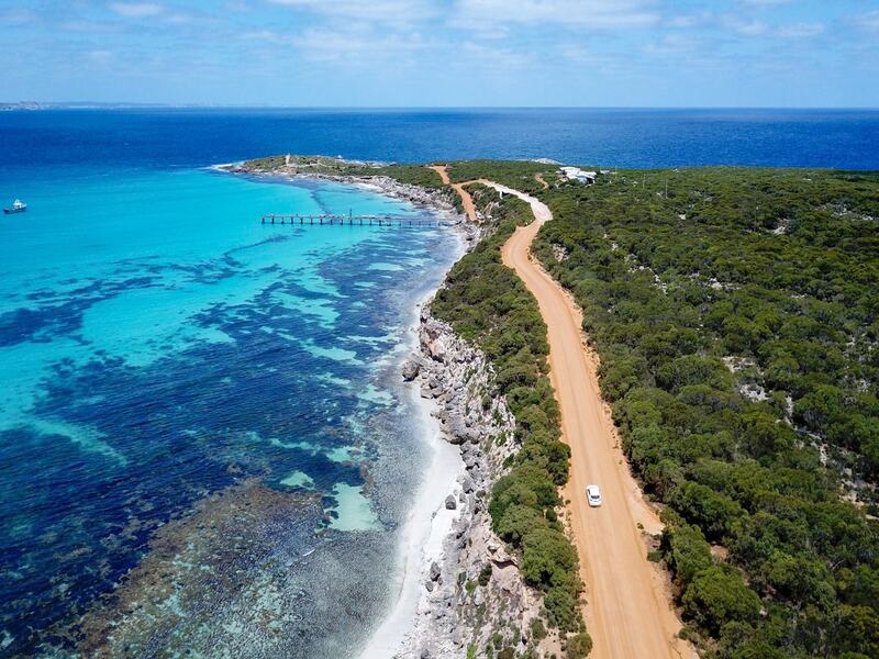 An aerial view of Vivonne Bay and Vivonne Bay camping ground on Kangaroo island. The crystal clear blue water of the bay is to the left of the image, while a dusty orange dirt road seperates the ocean from the green bushland to the left. the campground can be seen in the distance toward the background of the image