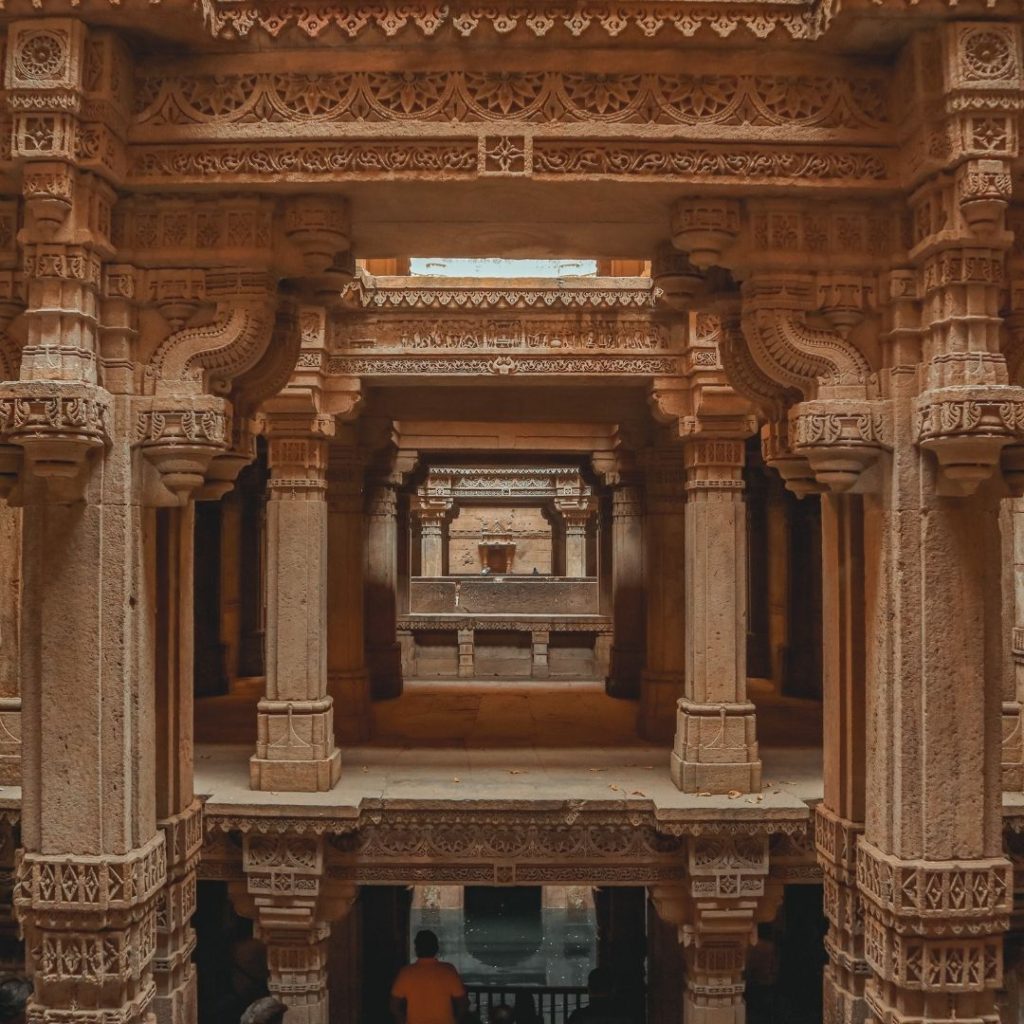 an internal image of the stepwell. Huge pillars with ornate carvings stretch from floor to ceiling. A top each pillar from left to right are more carved ceilings. A small amount of the stepwell water is visible at the very bottom of the image and a man in an orange shit is standing behind a guard rail admiring the structure