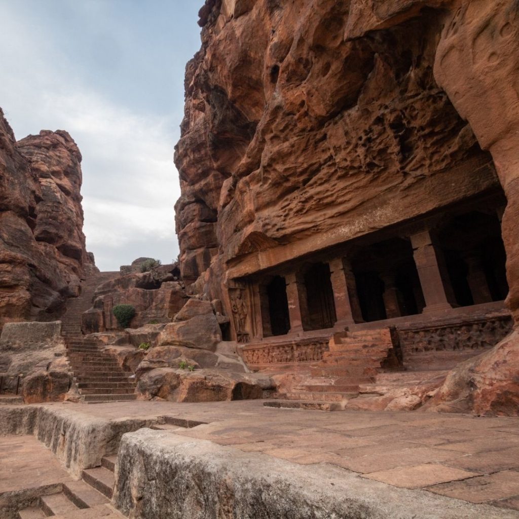 a side on view of the four carved out caves on the right of the image and some of the many steps needed to climb to the caves can be seen on the left of the image. The caves have been carved out of a natural red rock in the mountain