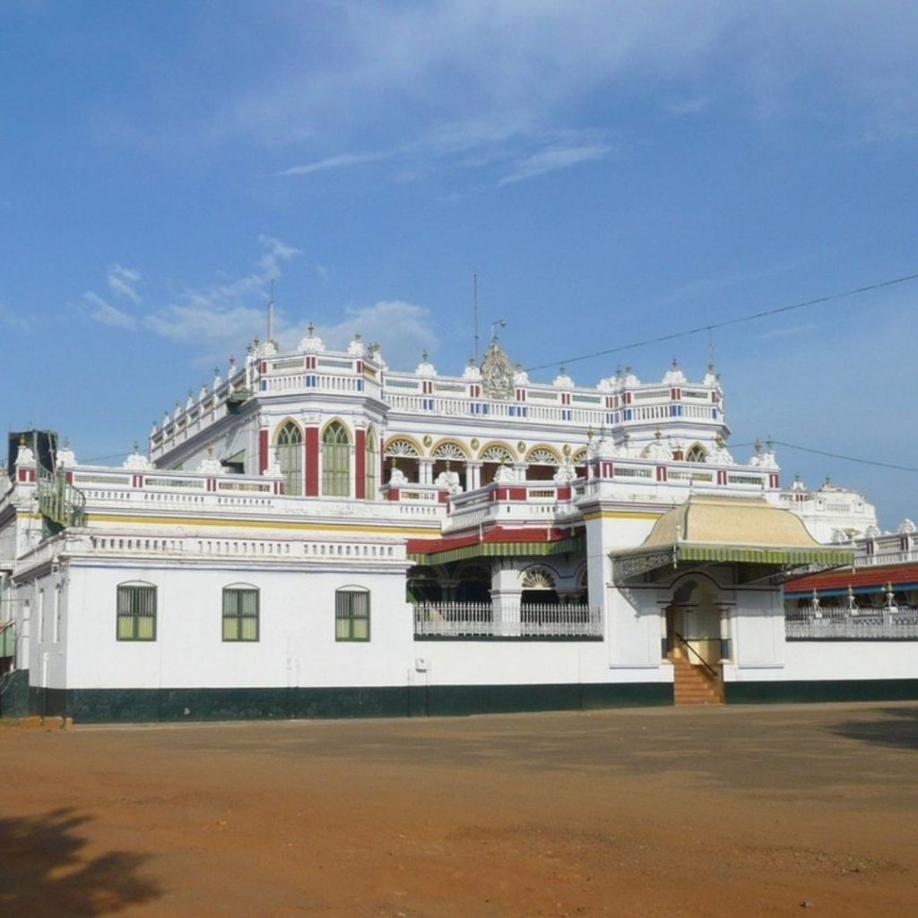 a slightly side on view point of Chettinad Palace. It is a white building with green, red and gold accents in the windows, doorways and arches