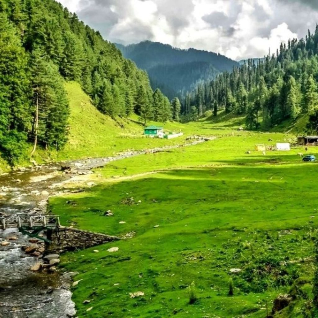 A natural landmark in India's Kasmir region. A lush green low lying valley with tree covered hills stretching upward left and right of the image, in the back ground are more large tree covered mountains. The grey and white clouds cover every inch of the sky. to the left of the image a steam meanders from the background to the foreground. A stone and wooden bridges crosses the water. There is a small green hut to the left in the background and a brown wooden hut to the right in the middle ground of the image