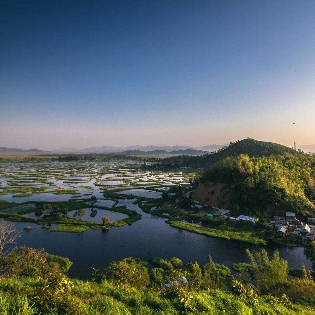 an aerial view of India's largest natural fresh water lake from a top a hill. in the middle of the lake to the right of the image is a big hill with many buildings at the base, Stretching from the hill all the way to the left of the image is the water separated by little stretches of green land and some trees.