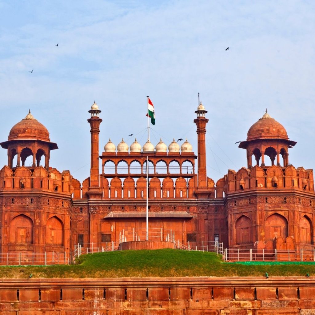 a front on image of the Red Fort. The Indian flag flies between two minarets in the centre of the image and several birds fly high above the towers in the blue sky