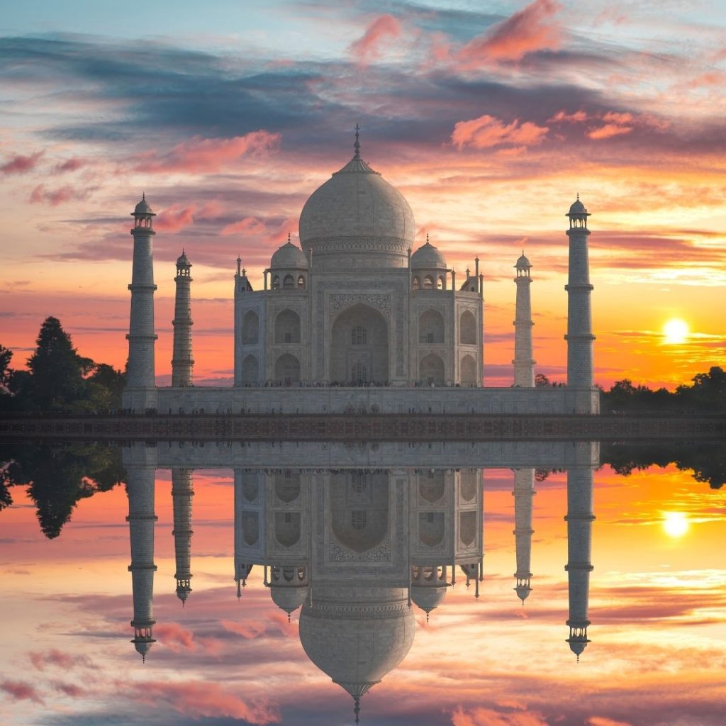 The most famous of India's landmarks. an image of the Taj Mahal at sunset. the sky is a dusty pink with shades of blue sky and the orange sun setting on the right of the image. The entire scene is mirrored perfectly in the lake in front of the Taj Mahal