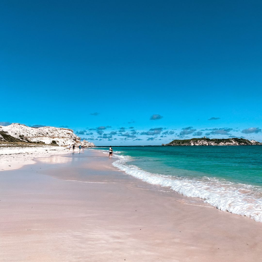 a view along the waters edge of Hamelin Bay beach looking towards the cliffs.. The white cliffs are on the left of the image and the ocean to the right. In the water is an island. On the white sandy beach are a few people walking in the water