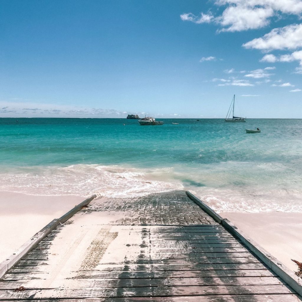 the Hamelin Bay boat ramp leading down the centre of the image into the clear blue water. there is white sand either side of the boat ramp. In the water are a few boats against the blue sky. there a few white fluffy clouds top right of the image